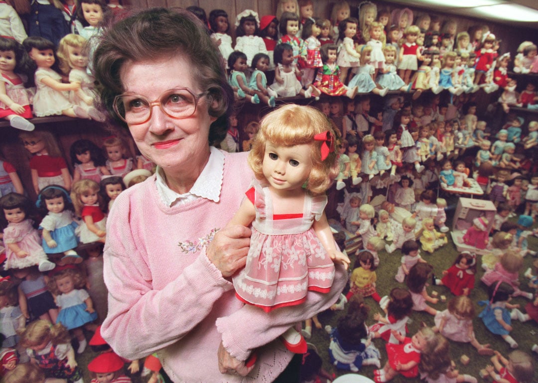 Ruth Kibbons holds the first Chatty Cathy doll she bought, a 1959 model, that started her 500-plus collection of the dolls, which are displayed behind her.