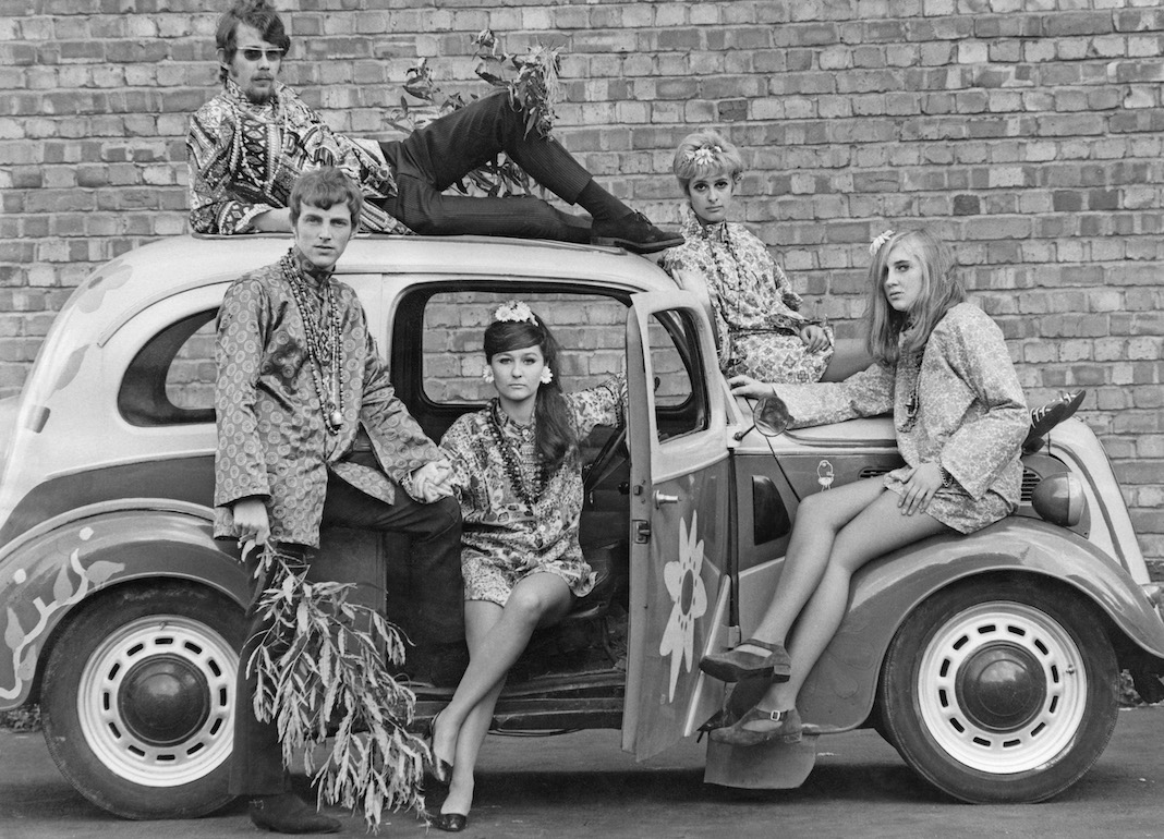 Young people pose in and on a car while modeling factory-made versions of Flower Power clothing.
