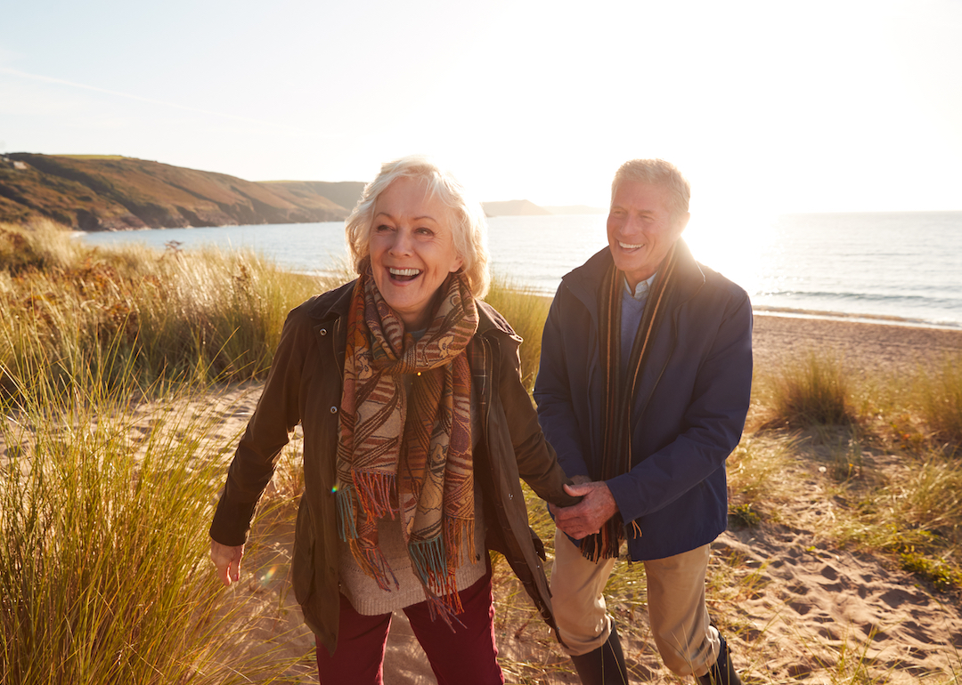 Retired couple smiling and laughing while walking on sand dunes in chilly weather.