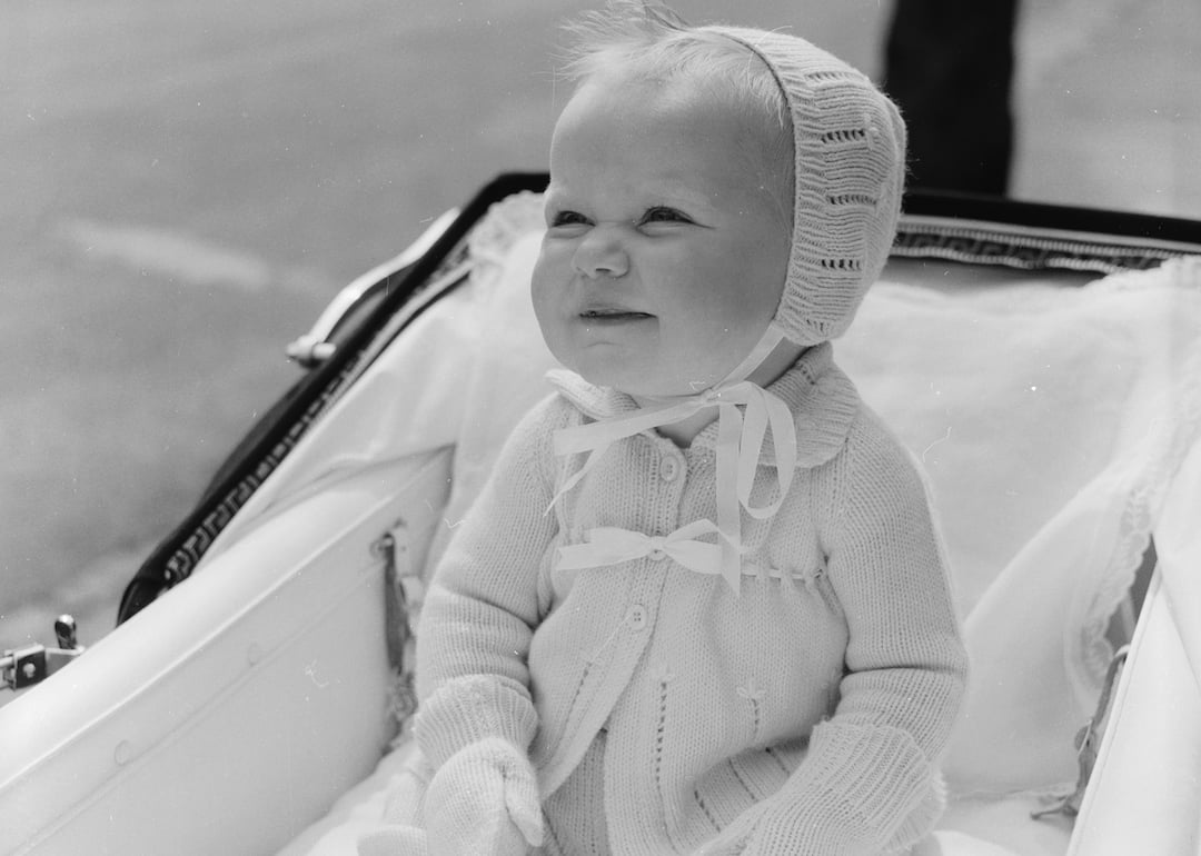 An infant, snug in matching mittens, bonnet and woolen booties, smiles in their carriage.