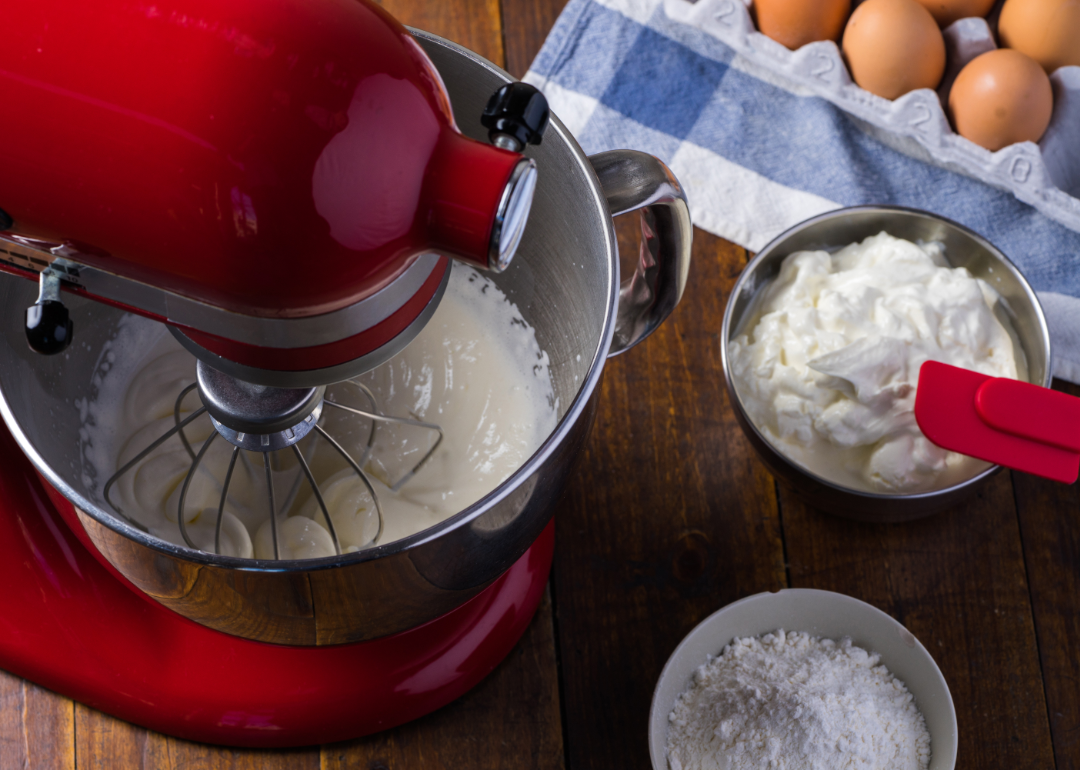 A red mixer with eggs, flour, sugar and cream on a blue and white tablecloth.