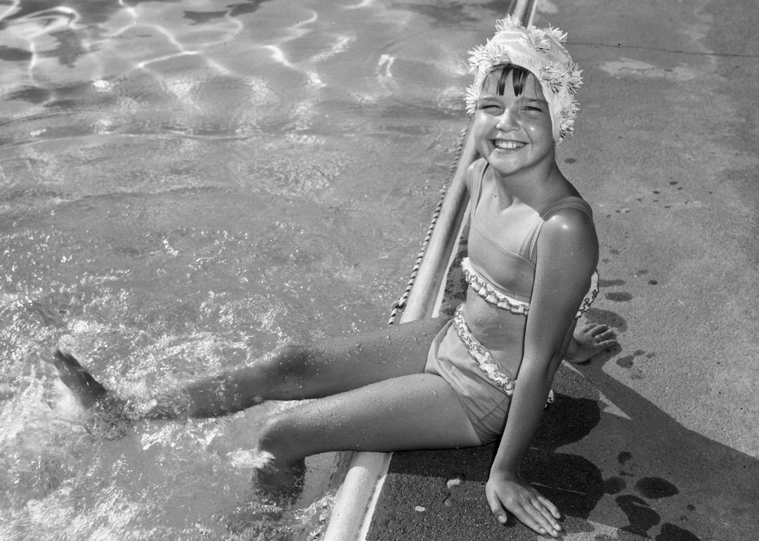 A young child in a swim cap and a two-piece bathing suit sits on the edge of a swimming pool and splashes their feet in the water in the 1960s.