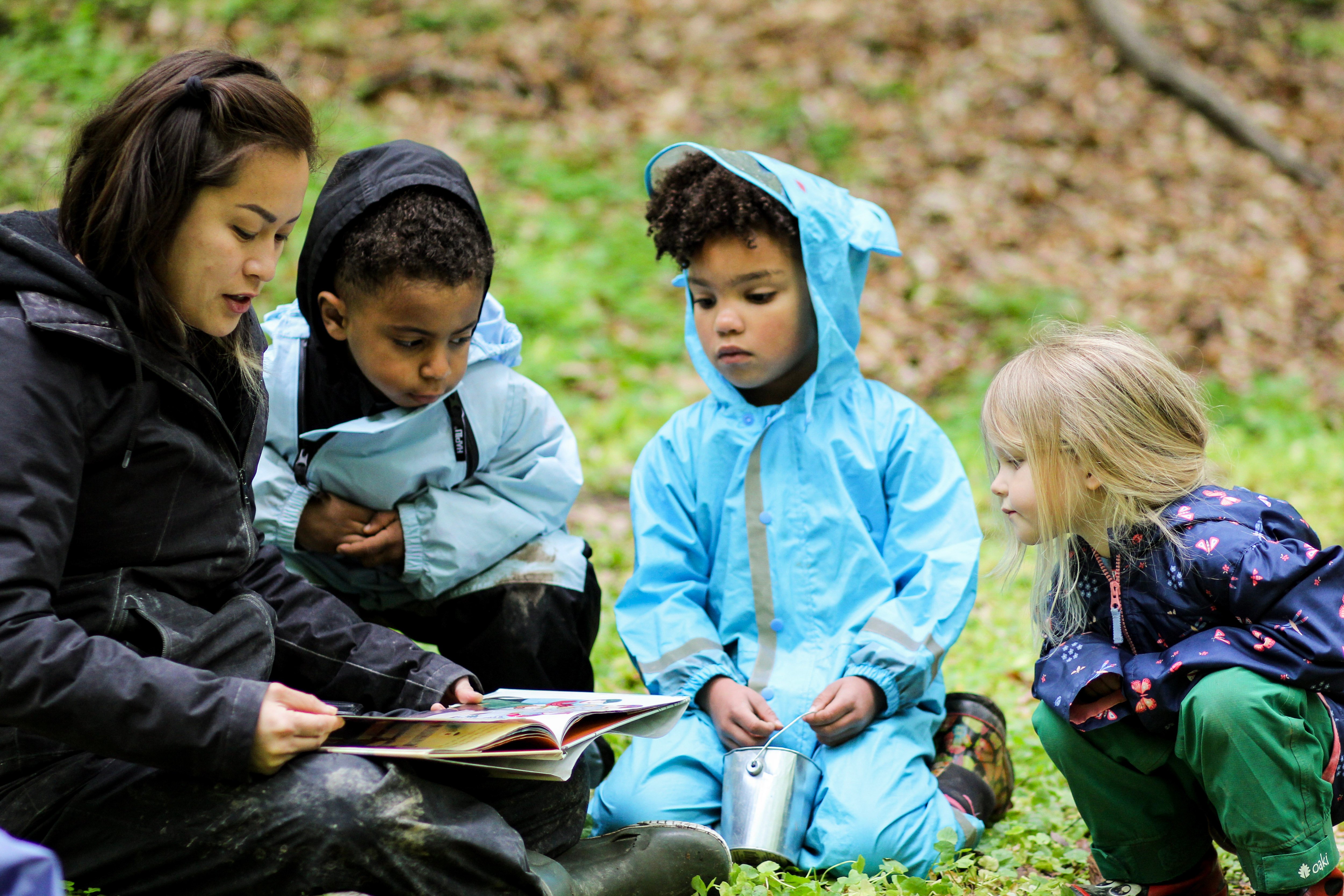 An adult sits with three children outside, holding a book and showing the children the pages.