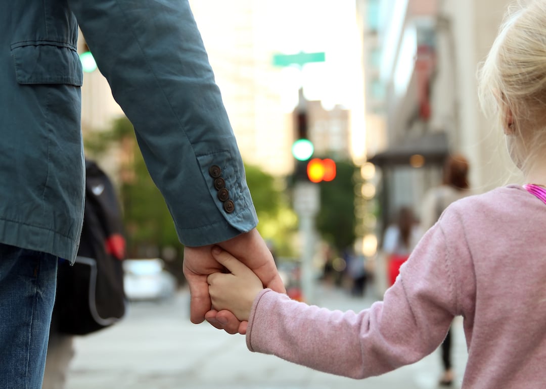 Parent holds child's hands in a city as they wait to cross the street.