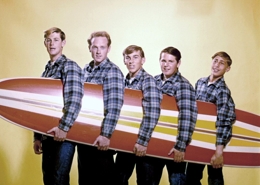 Rock and roll band "The Beach Boys" pose for a portrait with a surfboard in August 1962 in Los Angeles, California. (L-R) Brian Wilson, Mike Love, Dennis Wilson, Carl Wilson, David Marks. 