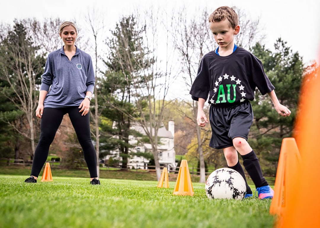 Kid kicking a soccer ball while his private coach observes.