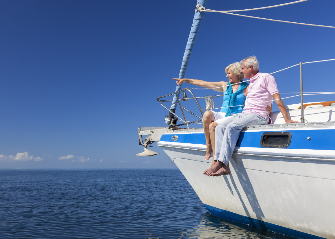 A happy senior couple sitting on the side of a sail boat on a calm blue sea looking and pointing to a clear horizon