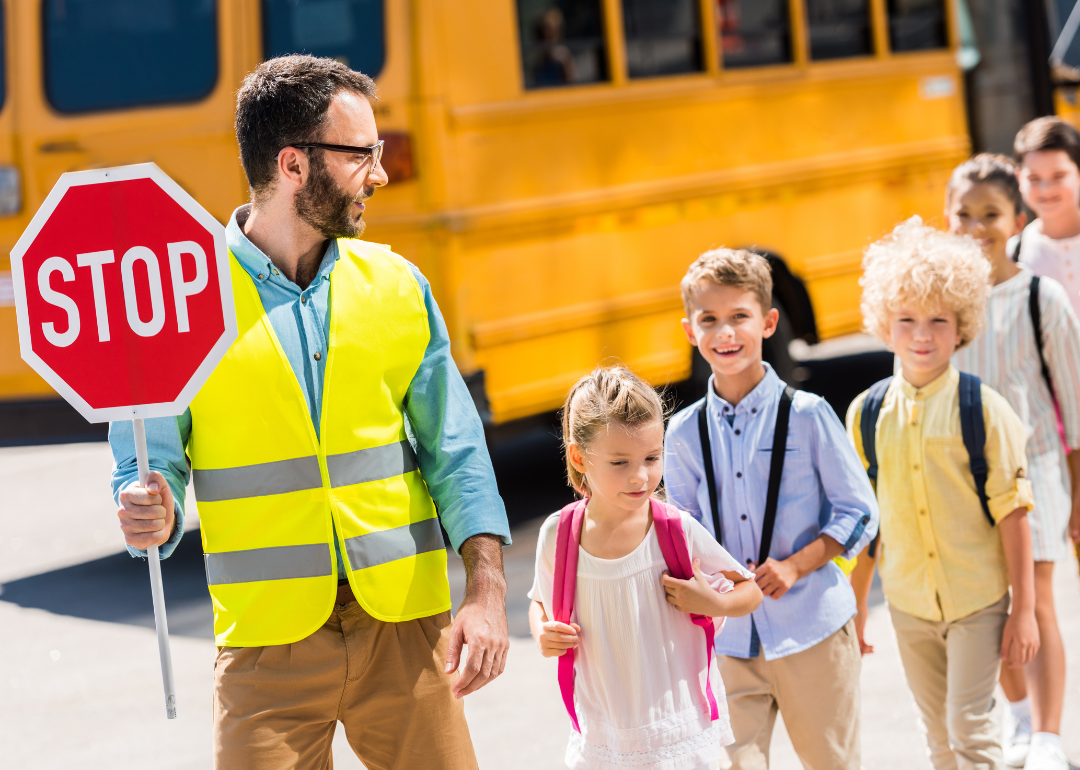 Traffic guard helping young school children cross the road in front of a school bus