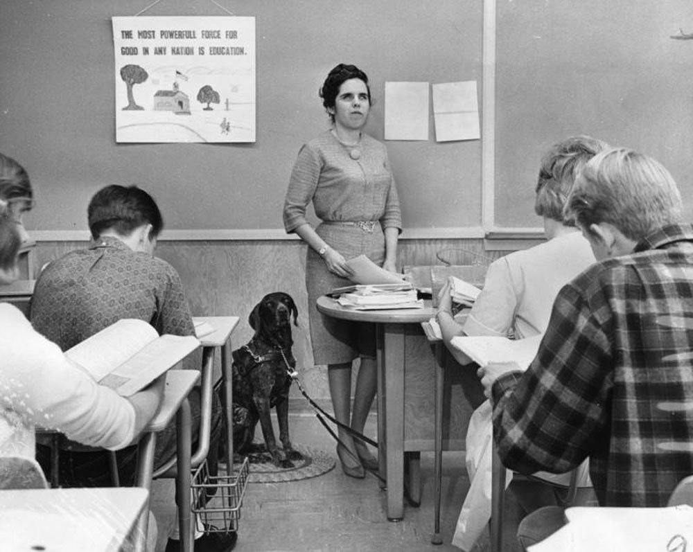 Black and white photo of a blind teacher and her dog in front of a class