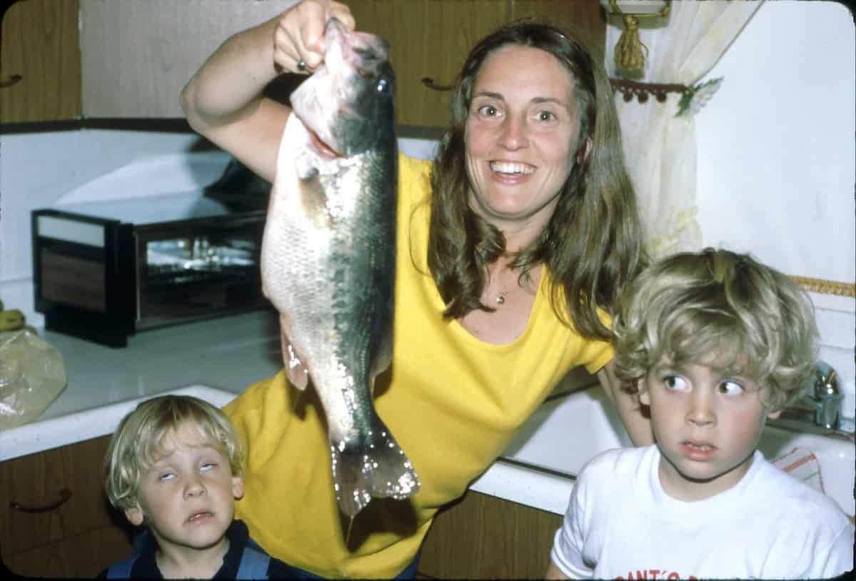 Mom smiling while holding up a fish in a kitchen, her two young boys have unpleasant expressions