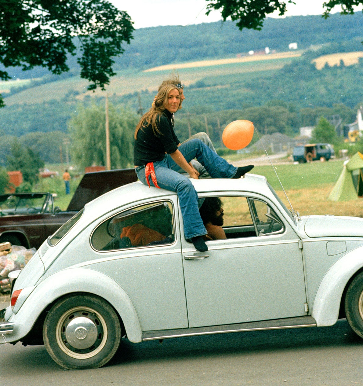 A couple sitting on the roof of a white VW bug driving down the road
