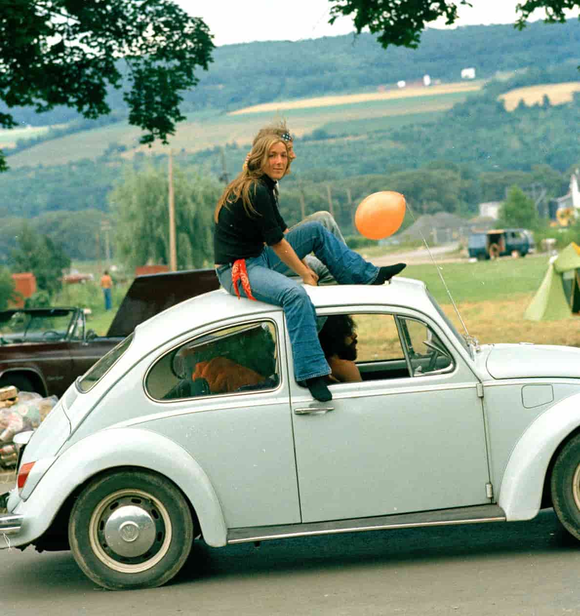 A couple sitting on the roof of a white VW bug driving down the road