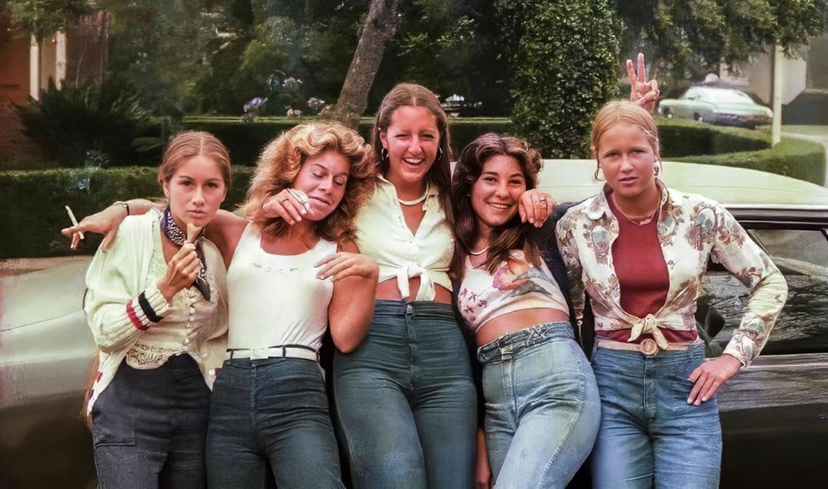 Color photo of five teenage girls leaning against a car on a tree-lined suburban street in San Diego in 1977. They wear high-waisted jeans and various casual tops including a tied blouse, a floral shirt, and a cardigan. One makes a peace sign and another rests her head on a friend's shoulder. Green hedges and parked cars are visible behind them.