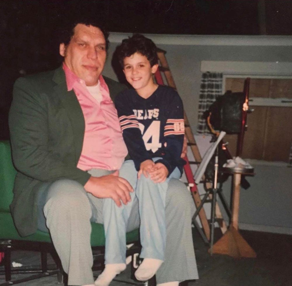 A large man in a green blazer and pink shirt sits with a young boy wearing a Chicago Bears jersey on what appears to be a film set, with lighting equipment visible in the background.