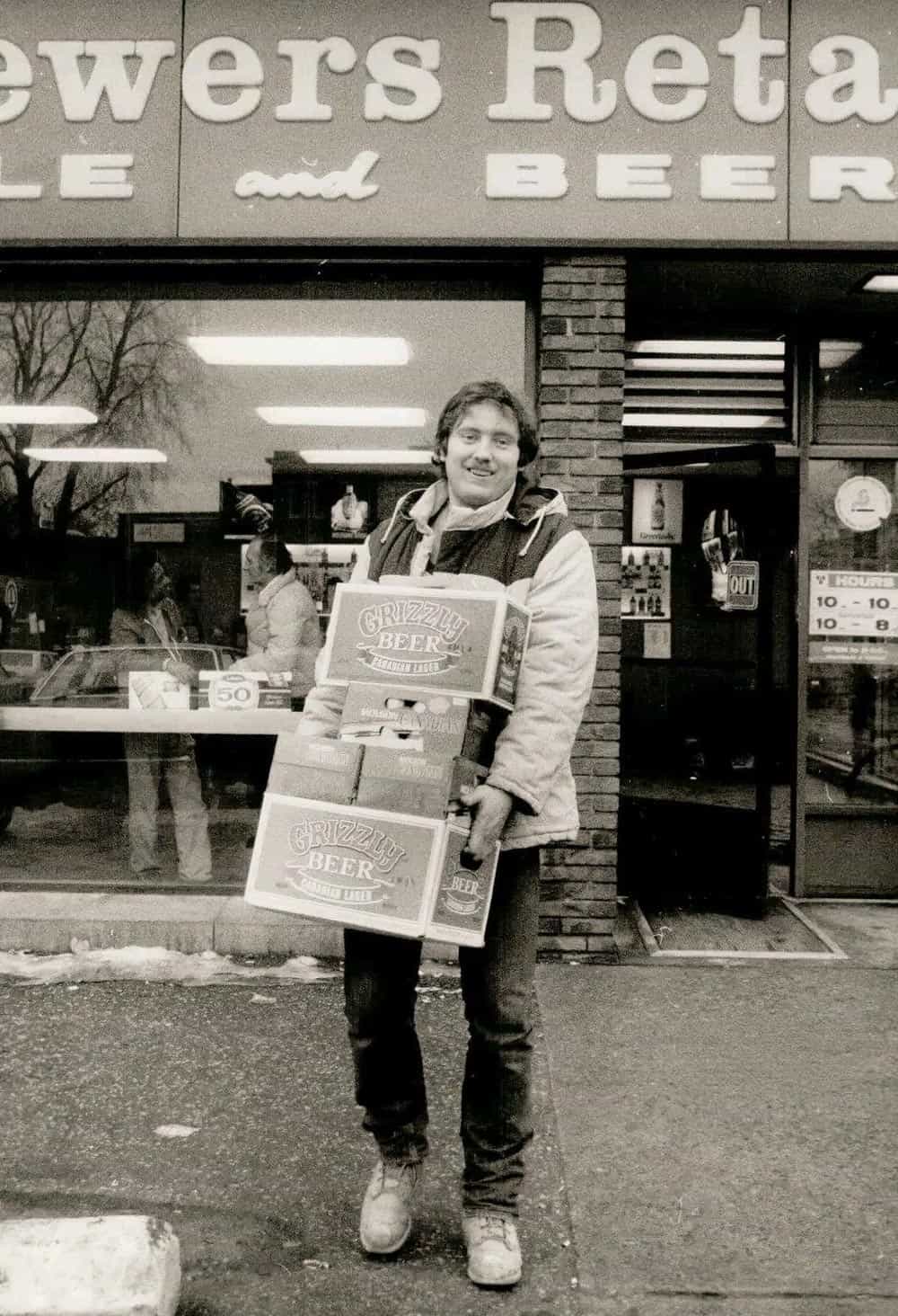 Black and white photo of a man carrying many boxes of beer leaving a Brewers Retail store