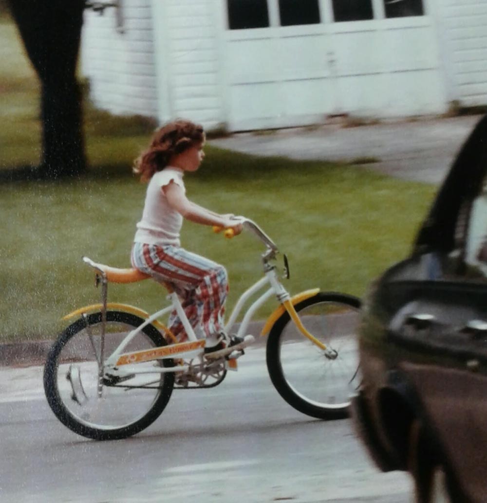 Color photo of a young girl with curly brown hair riding a yellow and white banana-seat bicycle down a suburban street in 1977. She wears a white t-shirt and red plaid shorts. A white house and green lawn are visible in the background.