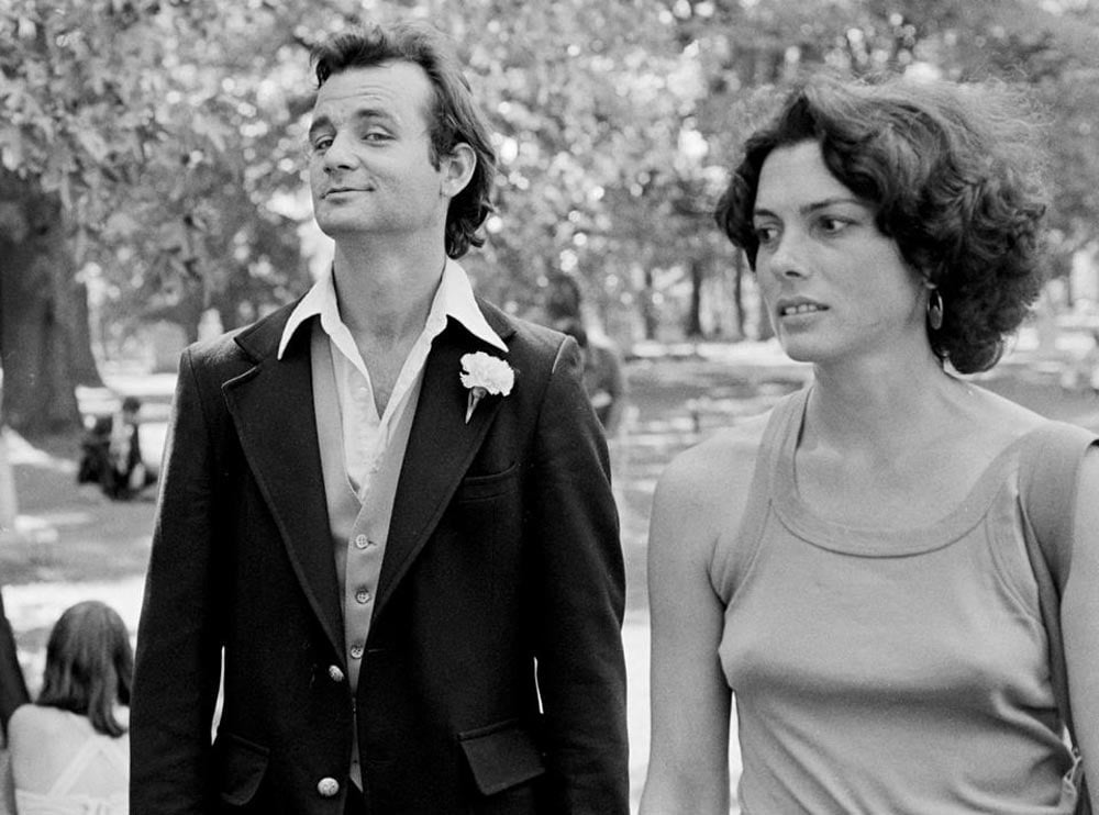 Black-and-white photo of Bill Murray and a woman walking outdoors in 1977. Murray wears a dark blazer over an open-collared shirt with a carnation boutonniere; the woman beside him wears a sleeveless top and hoop earrings. Trees are visible in the background.