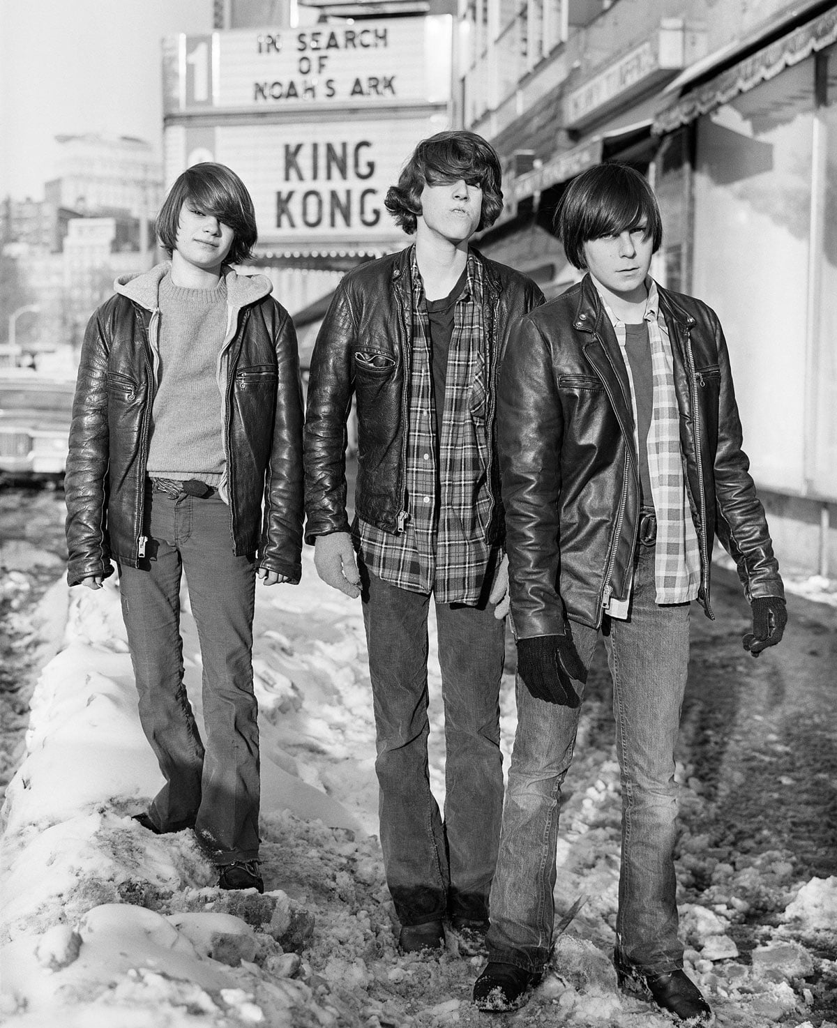 Black-and-white photo of three teenage boys in leather jackets walking down a snowy Boylston Street in Boston. All three have bowl-cut hair and wear plaid shirts underneath their jackets. A movie theater marquee behind them reads "King Kong" and "In Search of Noah's Ark."