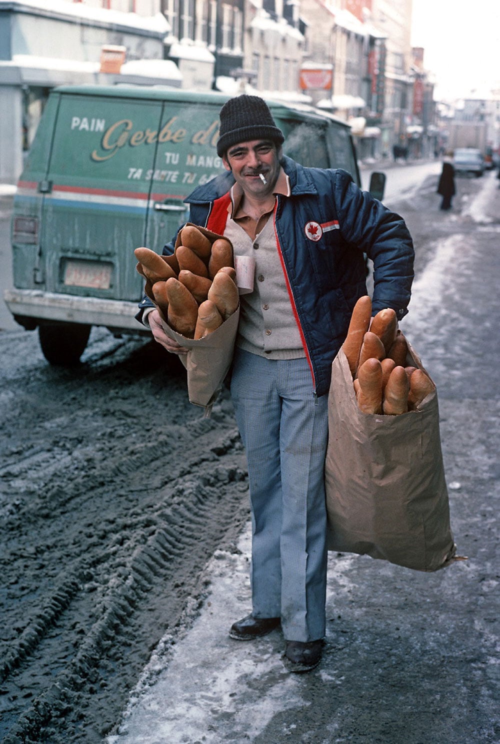 Color photo of a smiling bread deliveryman standing on a snowy street in Quebec City in 1977. He wears a dark knit cap and a blue jacket with a Canadian maple leaf patch, and holds two large paper bags stuffed with baguettes. A green Gerbe d'Or bread delivery van is parked behind him.