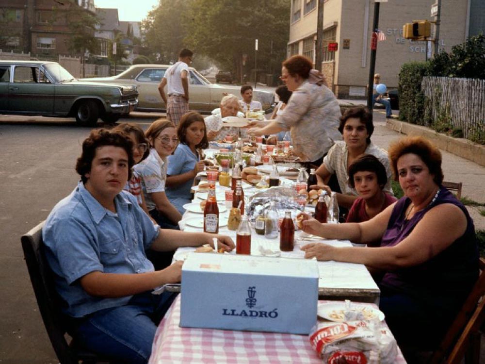 Color photo of a long outdoor table filled with food, condiments, and drinks at a Brooklyn block party in 1977. Roughly a dozen people of various ages sit along both sides of the table, which is covered with a pink checkered cloth and lined with ketchup bottles, soft drinks, and plates of food. A school building is visible in the background on a quiet residential street.