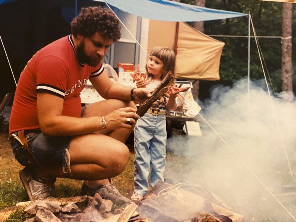 A bearded man crouches by a smoky campfire with a small girl in jeans and a Mickey Mouse belt, both holding sticks over the flames, with a tent and tarp visible behind them in the woods.