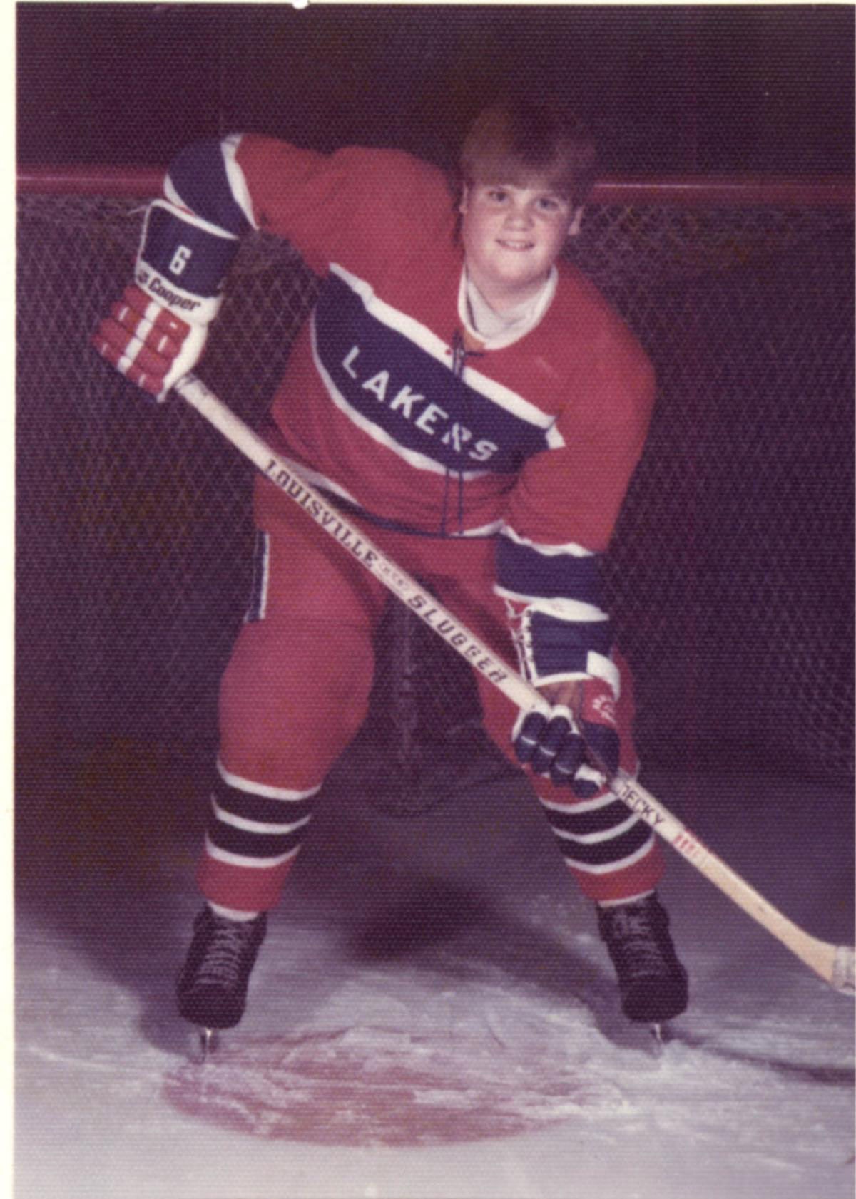 Color photo of a teenage Chris Farley posing in a red and blue Lakers youth hockey uniform, number 6, holding a Louisville Slugger hockey stick on the ice in Madison, Wisconsin, 1977. He grins broadly at the camera.