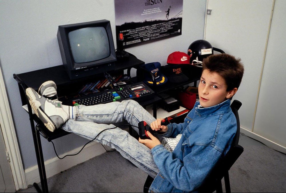 A young teenage boy in a denim jacket and acid wash jeans sits with his feet up on a desk, holding a joystick in front of a computer, with an Empire of the Sun movie poster visible on the wall behind him.