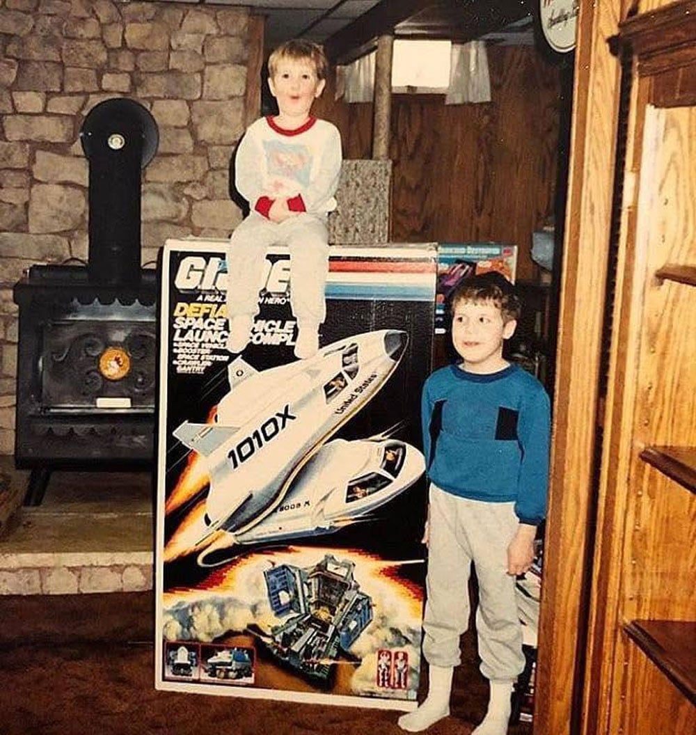 Two young boys in pajamas and sweats pose next to an enormous G.I. Joe Defiant Space Vehicle Launch Complex box on Christmas morning in a wood-paneled living room with a cast iron wood stove.