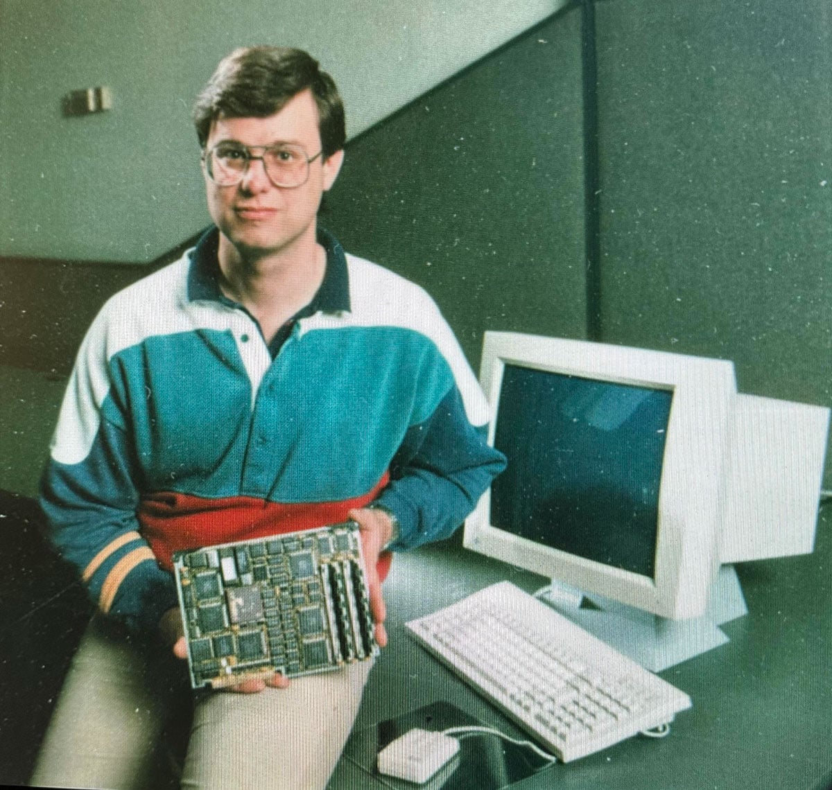 A man in a color-block polo shirt sits next to a desktop computer and monitor, holding a circuit board toward the camera in what appears to be a promotional or magazine photograph from the late 1980s.