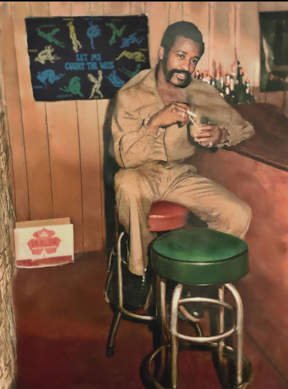 Color photo of a man seated on a red barstool at a wood-paneled basement bar in Philadelphia, 1977. He wears a tan jumpsuit and holds a drink, leaning back with one foot resting on a green stool beside him. A novelty poster hangs on the wall behind him and a case of beer sits on the floor to his left.
