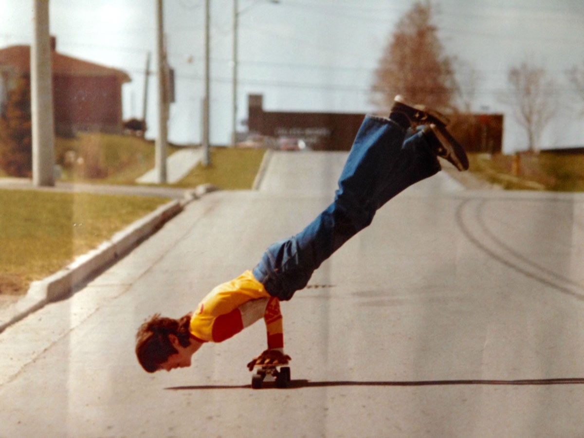 Color photo of a young person performing a handstand on a skateboard while rolling down a suburban street in 1977. They wear a yellow and red jersey and flared jeans, with their legs extended straight up in the air. A quiet residential neighborhood with utility poles and a brick building is visible in the background.