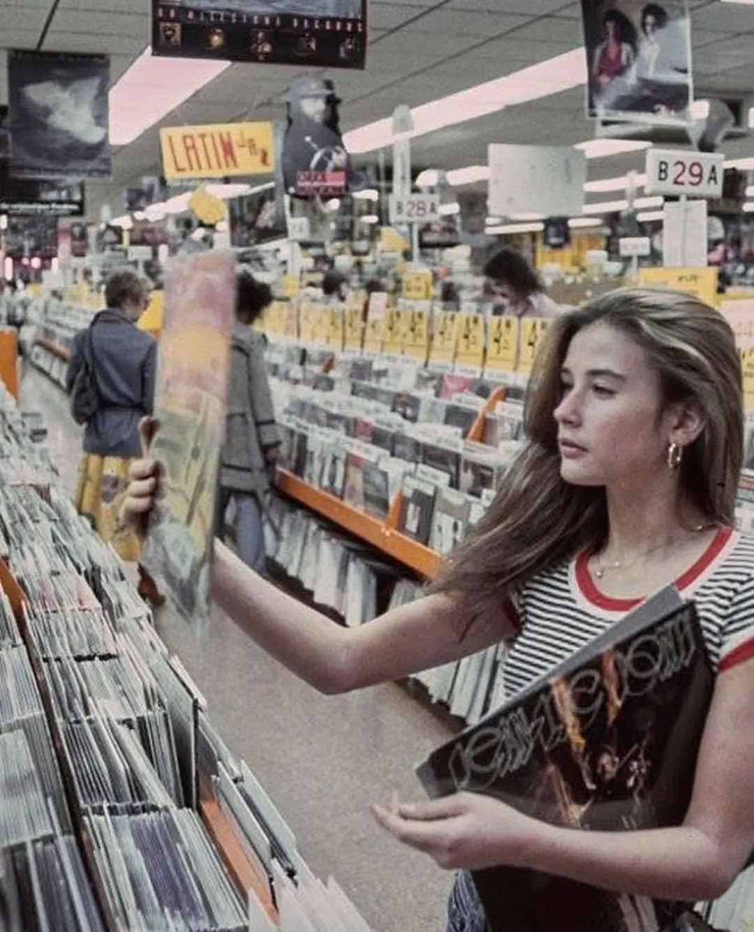 Color photo of a teenage Demi Moore browsing vinyl records at Tower Records on the Sunset Strip in Los Angeles, 1977. She wears a striped red-and-white tank top and hoop earrings, and holds a record album while standing in an aisle lined floor to ceiling with LP covers. A "Latin" section sign is visible in the background.