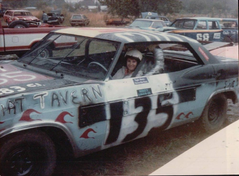A smiling woman behind the wheel of a demolition derby car