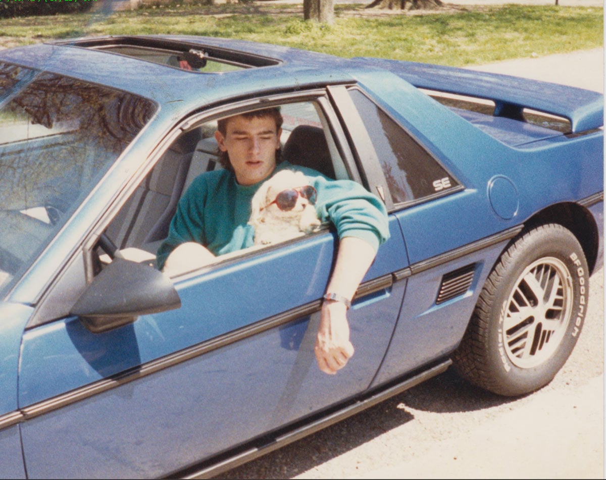 A young man in a teal sweatshirt sits in a blue 1984 Pontiac Fiero SE with a small white dog wearing red sunglasses perched on his lap, both looking out the open window.