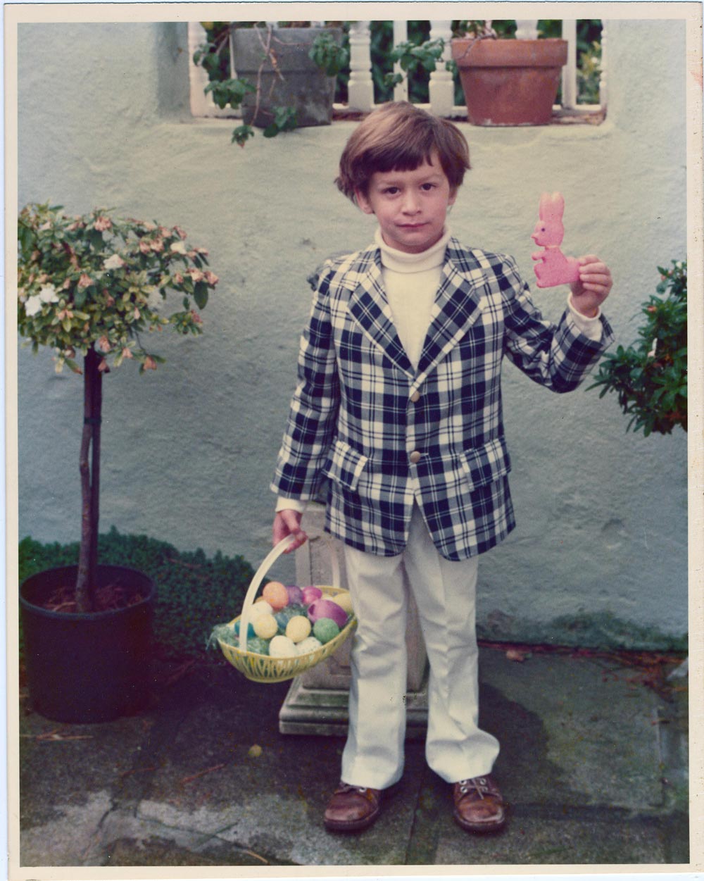 Color photo of a young boy standing in a garden courtyard in California on Easter, 1977. He wears a plaid blazer over a white turtleneck with white trousers and brown shoes, holds a yellow Easter basket full of colored eggs in one hand, and raises a pink sugar bunny cookie in the other. Potted topiary trees and a stucco wall are visible behind him.