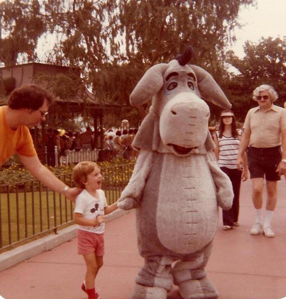 Color photo of a young toddler in a Mickey Mouse shirt and red shoes holding hands with the Eeyore character at Walt Disney World in 1977. The child's mouth is wide open in delight. A father in an orange shirt leans in from the left, and two adults walk past in the background.