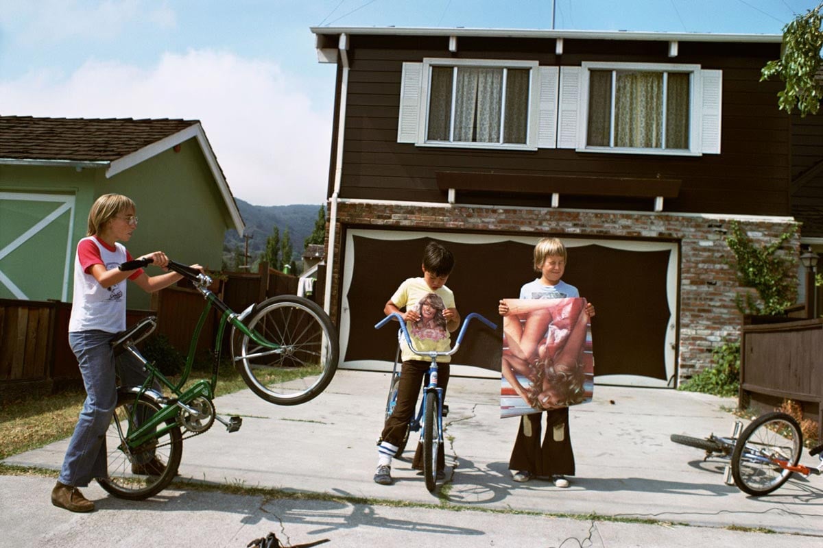 Color photo of three children standing with their bikes in a suburban driveway in California in 1977. The child on the left holds up a green bike's front wheel; the middle child straddles a blue bike; the child on the right holds a large Farrah Fawcett poster. A two-story brown house with a two-car garage stands behind them.