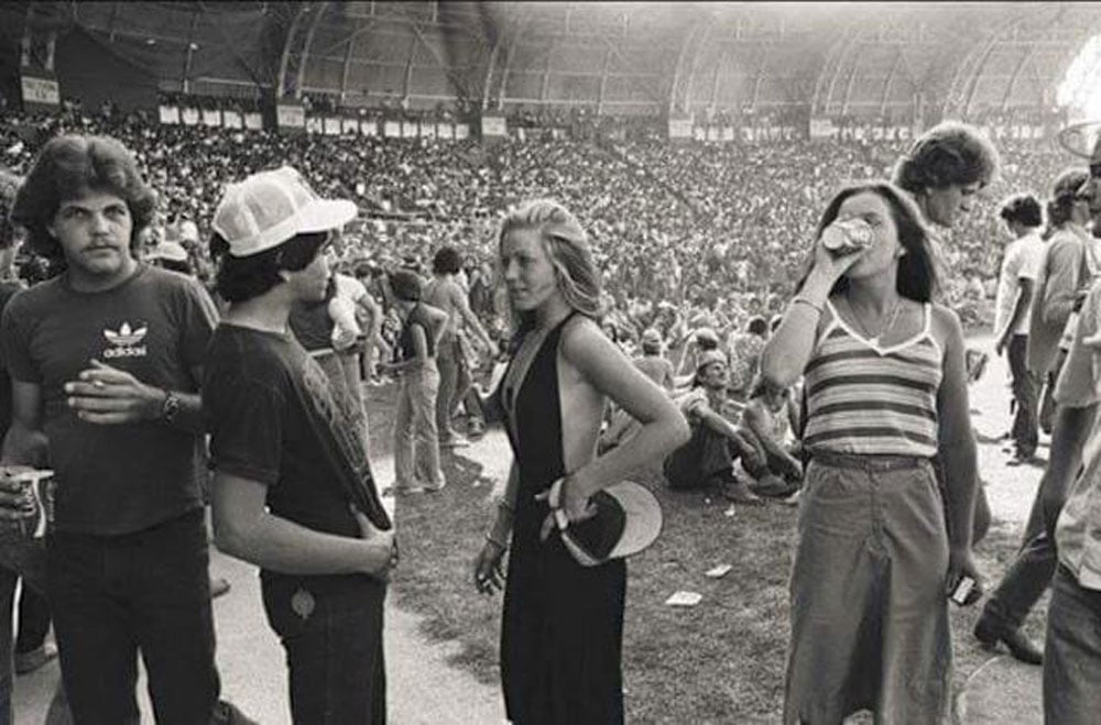Black-and-white photo of concertgoers at a packed indoor arena in 1977. In the foreground, a young man in an Adidas t-shirt and white cap faces a woman in a deep V-neck halter dress; a second woman in a striped tank top drinks from a can beside them. Thousands of fans fill the stadium behind them.