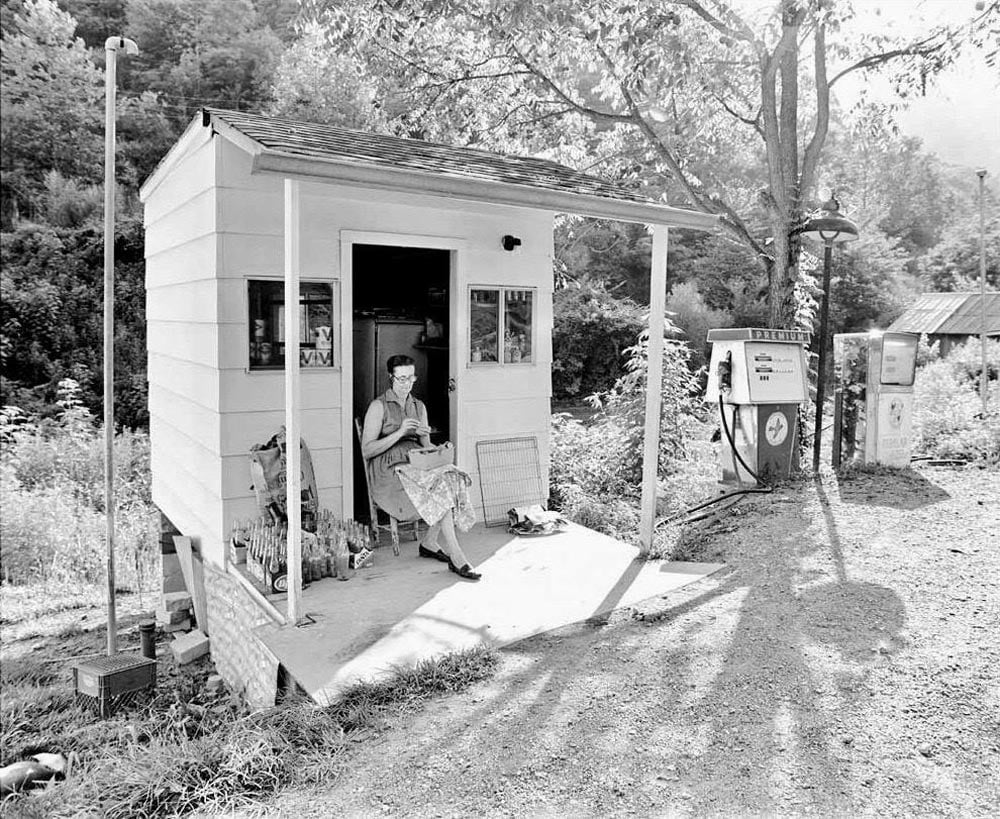 Black-and-white photo of a woman seated in the open doorway of a small white clapboard gas station in Knott County, Kentucky, in 1977. She sews in her lap while two vintage gas pumps stand to her right on a dirt lot surrounded by trees and overgrowth.