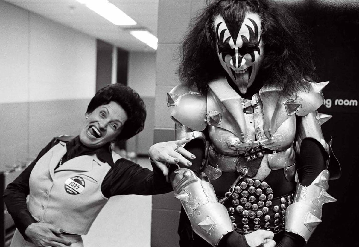 Black-and-white photo of Gene Simmons in full KISS Demon costume and makeup standing backstage with his mother Florence in 1977. Florence wears a vest with a KISS button pinned to it and laughs openly at the camera. Gene, in spiked armor and black and white face paint, stands beside her grinning.