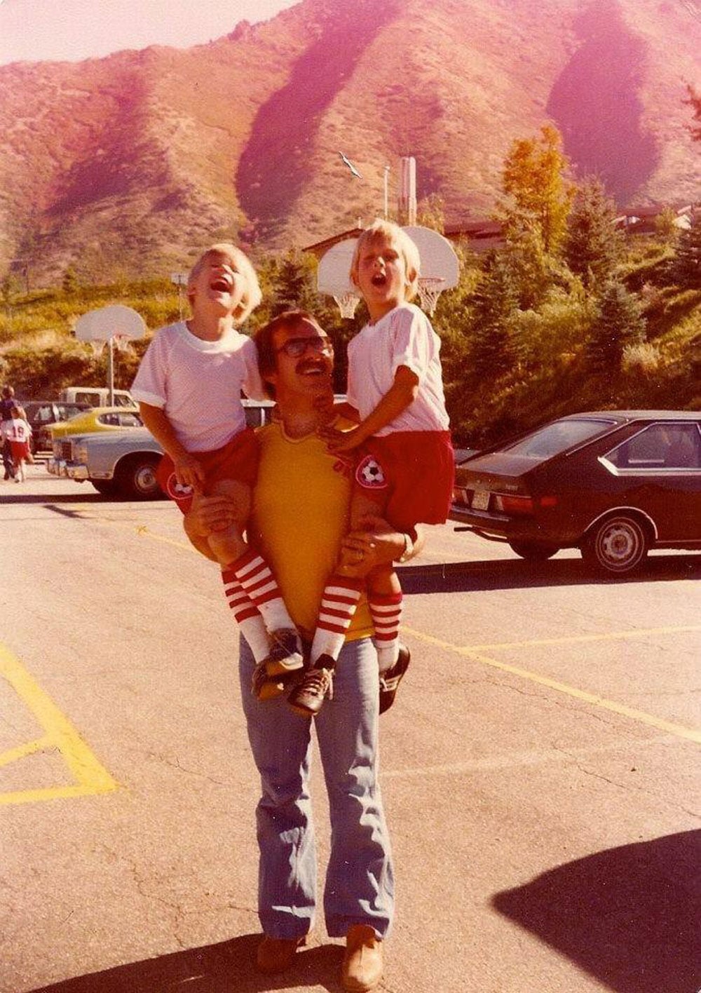 Color photo of a father holding two young blond boys in soccer uniforms, one on each arm, in a parking lot in 1977. All three are laughing with their heads thrown back. Red and brown mountains rise in the background behind a basketball hoop and a row of parked cars.
