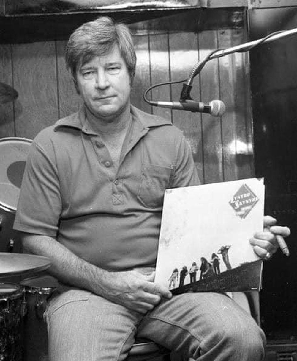 Black-and-white photo of Leonard Skinner, a middle-aged man with short hair, seated and holding a Lynyrd Skynyrd record album. A microphone stand and drum kit are visible behind him.