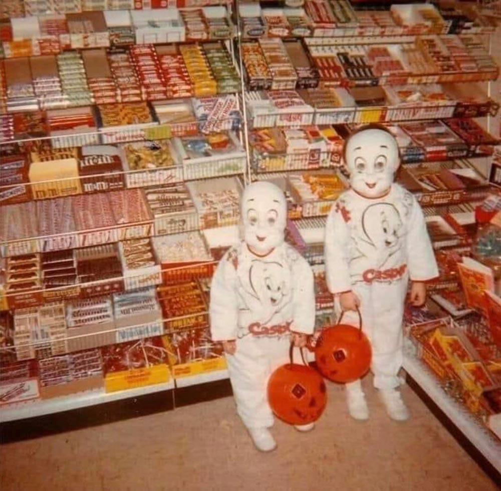 Color photo of two young children in matching Casper the Friendly Ghost Halloween costumes — white jumpsuits with Casper's face printed on the chest and molded plastic masks — each holding an orange plastic jack-o'-lantern candy bucket. They stand in front of floor-to-ceiling candy display shelves stocked with M&Ms, Reese's, and other candy bars.