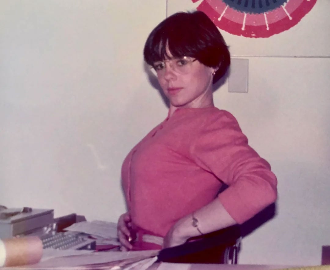 Color photo of a woman seated at an office desk in 1977, looking over her shoulder at the camera. She wears a pink top, wire-rimmed glasses, and a short dark bowl cut. Papers and a typewriter are visible on the desk; a red, white, and blue decorative bunting hangs on the wall behind her.