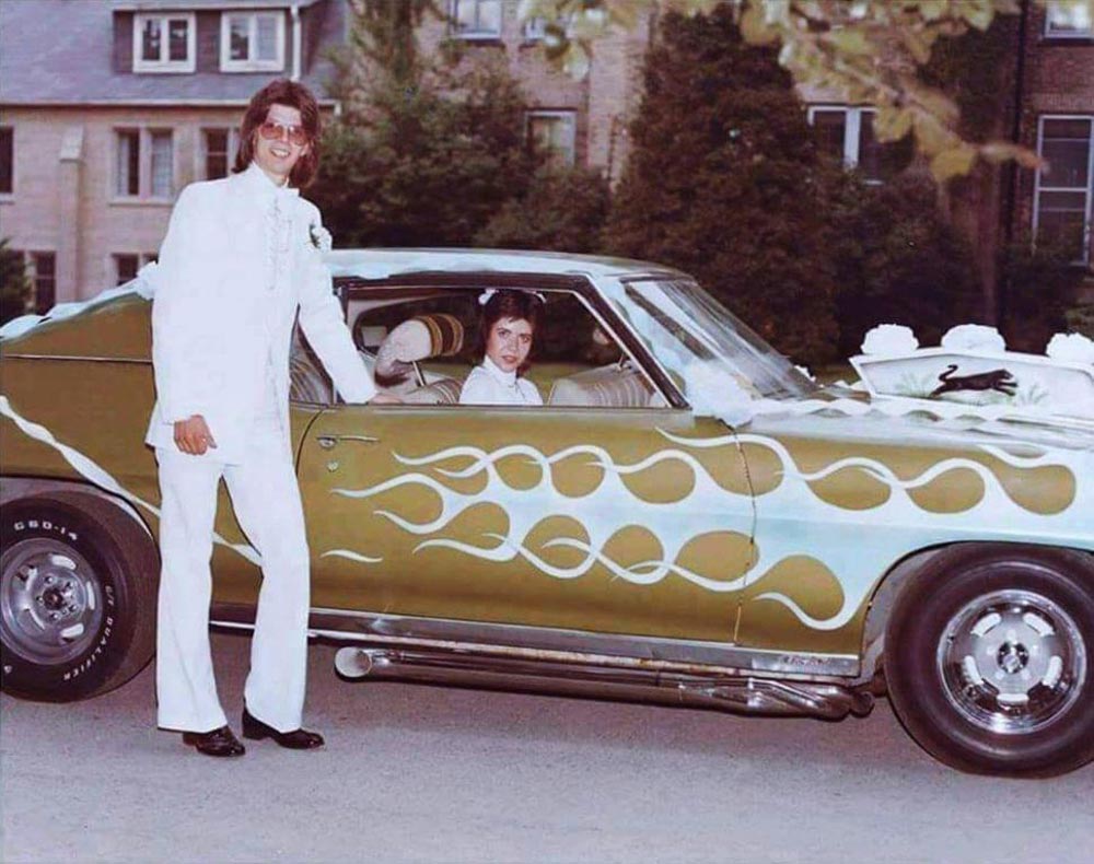 Color photo of a young groom in a white suit and aviator sunglasses standing beside a custom two-tone gold and light blue Corvette with white flame paintwork, decorated with white floral wedding ribbons. His bride sits in the passenger seat. A brick building and trees are visible in the background.
