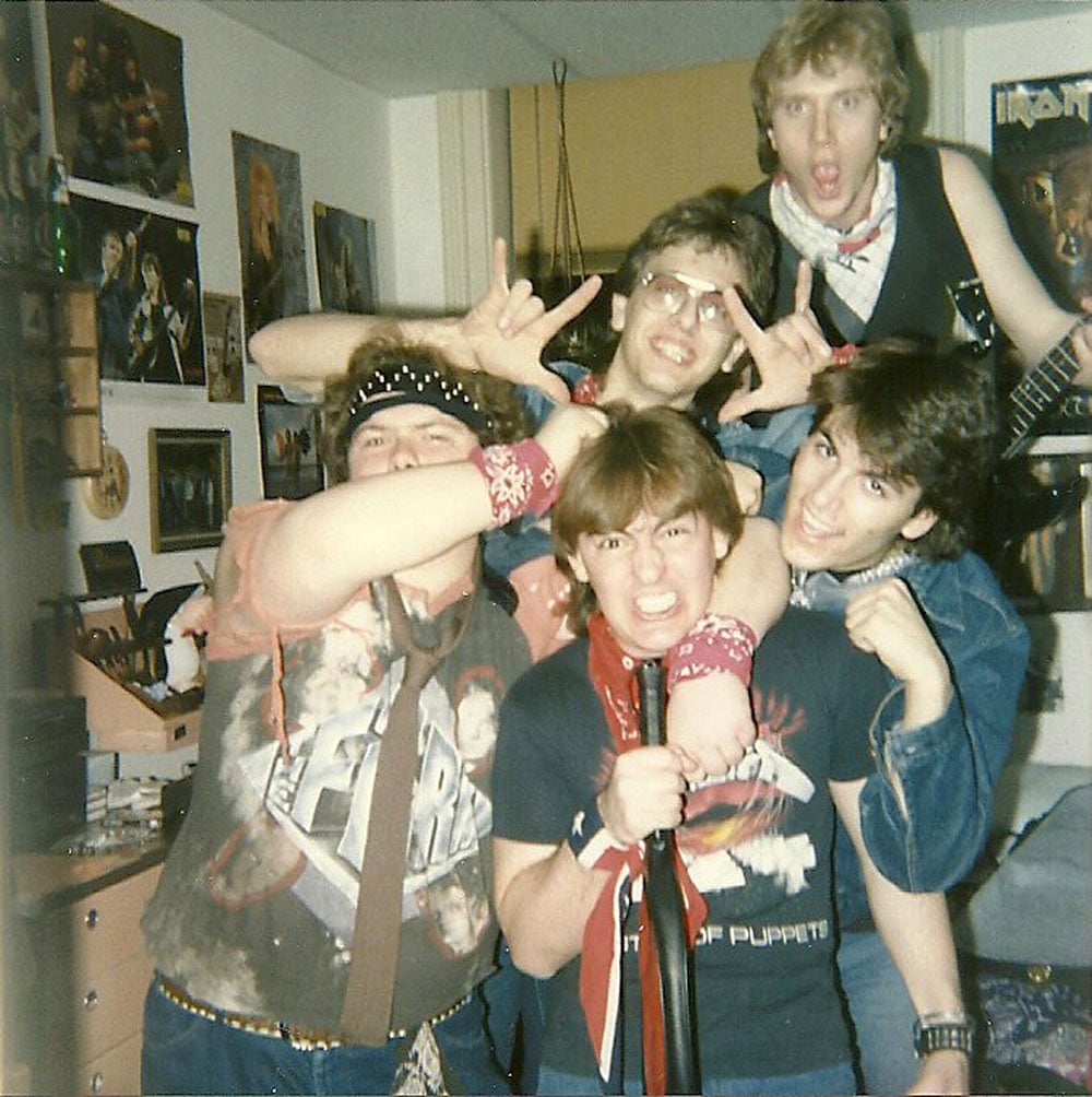 ive teenage boys crowd together in a bedroom covered in rock band posters, throwing up devil horns and mugging for the camera in band shirts, bandanas, and denim before a concert in 1987.