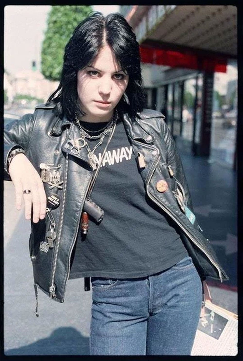 Color photo of a teenage Joan Jett standing on a street in Los Angeles in 1977. She wears a black leather jacket covered in pins and chains over a dark t-shirt, with jeans. Her black hair is cut short and she stares directly into the camera.