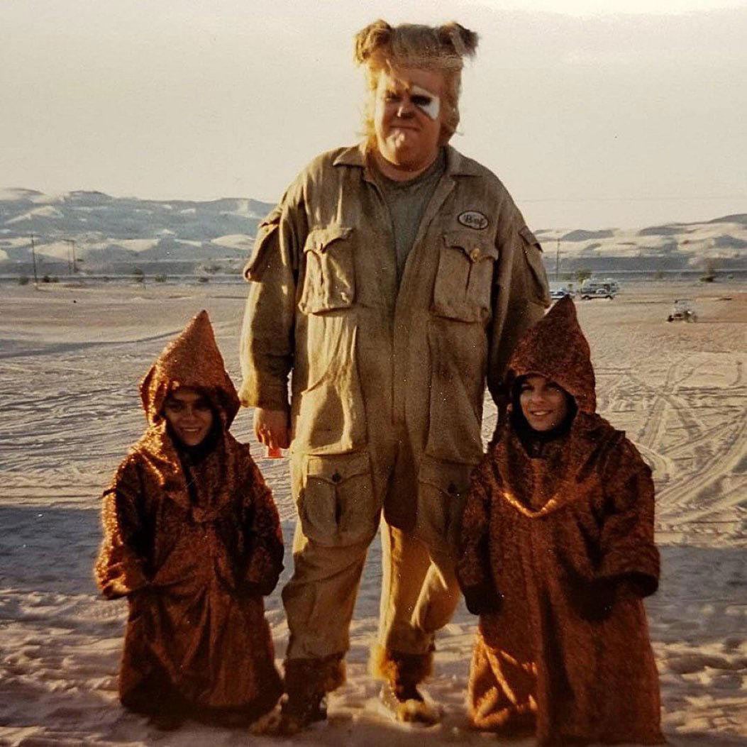 A large man in a khaki jumpsuit with a shaggy blonde wig and face paint stands in a desert landscape flanked by two small figures in hooded brown robes.