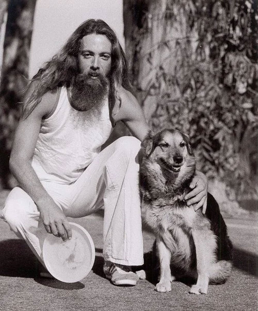Black-and-white photo of Ken Westerfield crouching outdoors beside a large mixed-breed dog in 1977. He has long hair and a full beard, wears a white tank top and white flared pants, and holds a Frisbee disc in one hand with his other arm around the dog.
