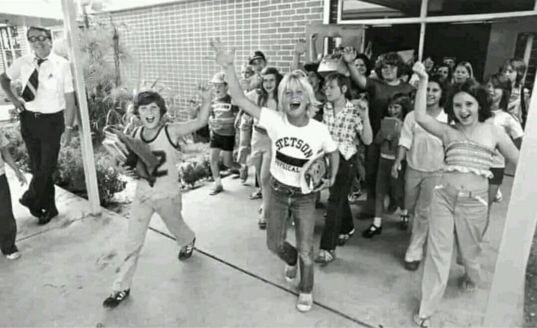 Black-and-white photo of a crowd of students streaming out of a school building on the last day of school in 1977. Three kids in the foreground cheer with their arms raised, one wearing a Stetson Physical Education t-shirt. A male administrator in a white shirt and tie watches from the left.